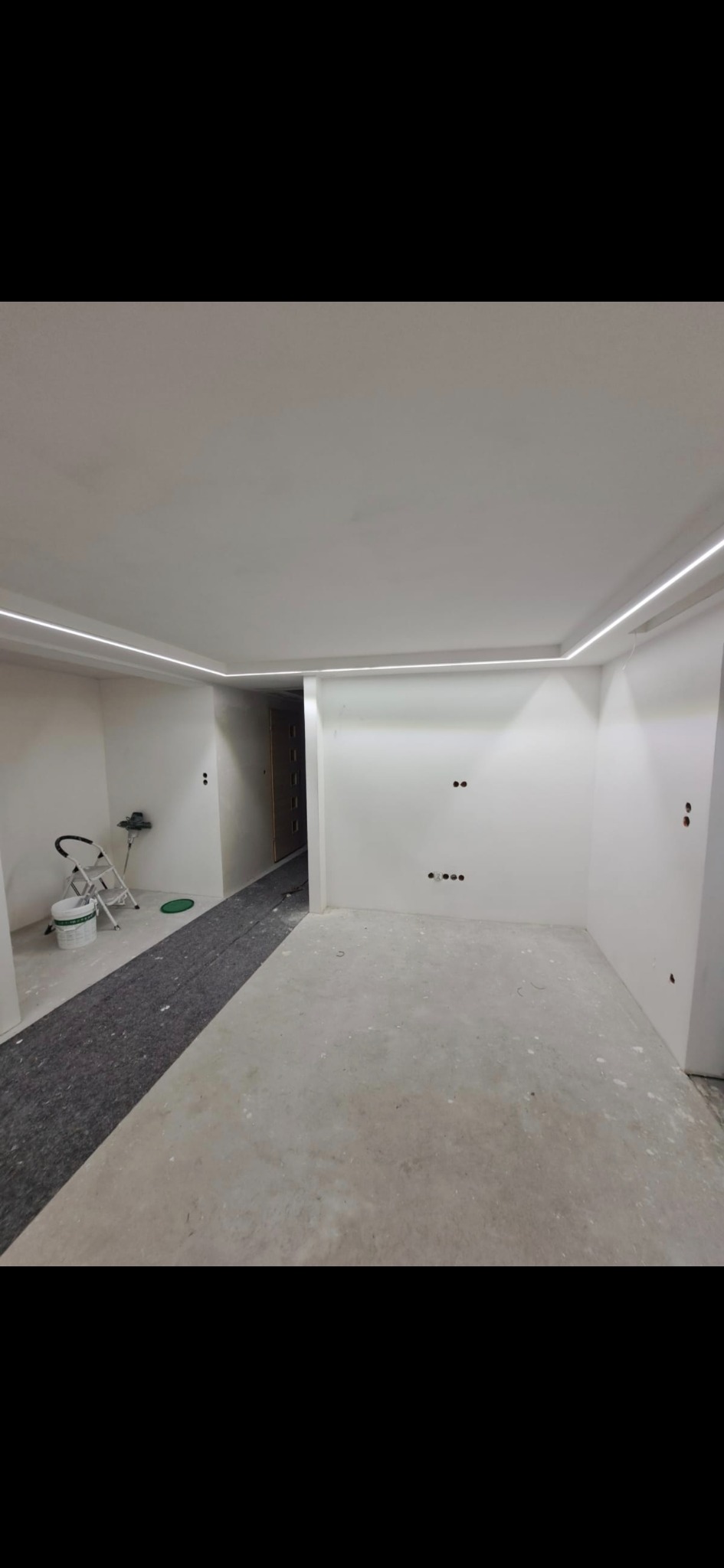 Wnętrze po remoncie: białe ściany z subtelnym oświetleniem LED przy suficie, przygotowane do dalszych prac wykończeniowych, widoczne zabezpieczenia podłogi.