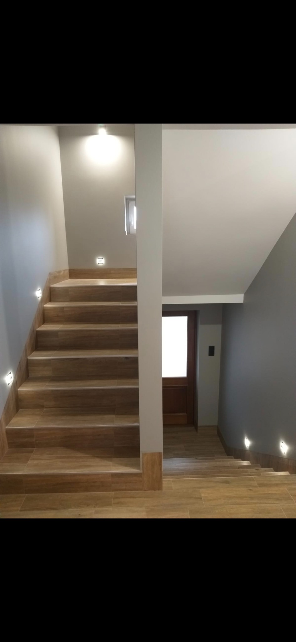 Wykończenie schodów z drewnopodobnych płytek, podświetlane stopnie, szara ściana i drzwi w tle. Nowoczesny design, oświetlenie LED.