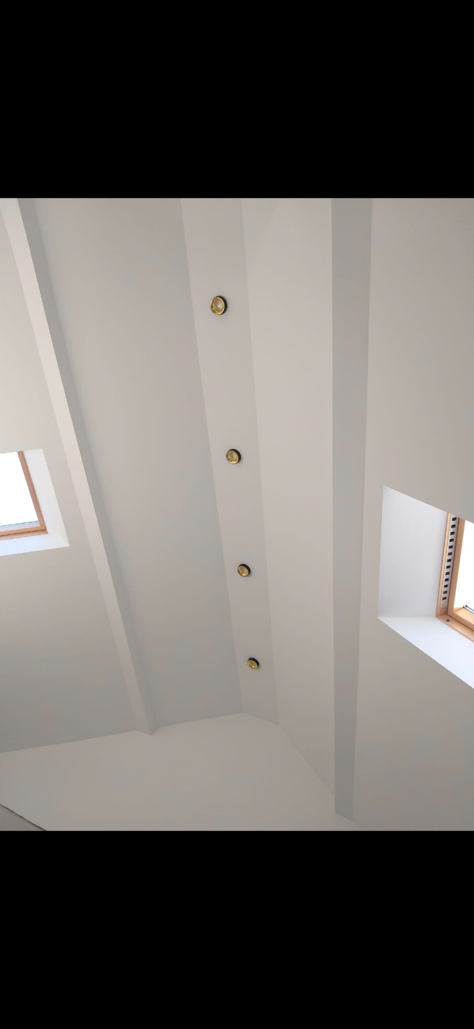 Białe, skośne poddasze z oknami dachowymi i rzędem punktowych lamp wzdłuż belki stropowej. Minimalistyczne wykończenie.