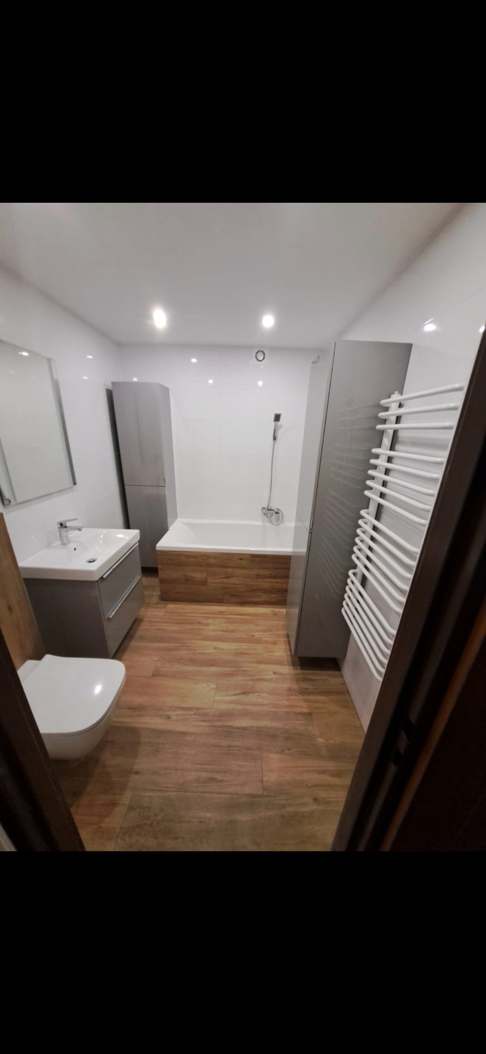 Nowoczesna łazienka po remoncie z drewnopodobną podłogą, białą armaturą i szarą zabudową meblową. Widok zza futryny drzwi.