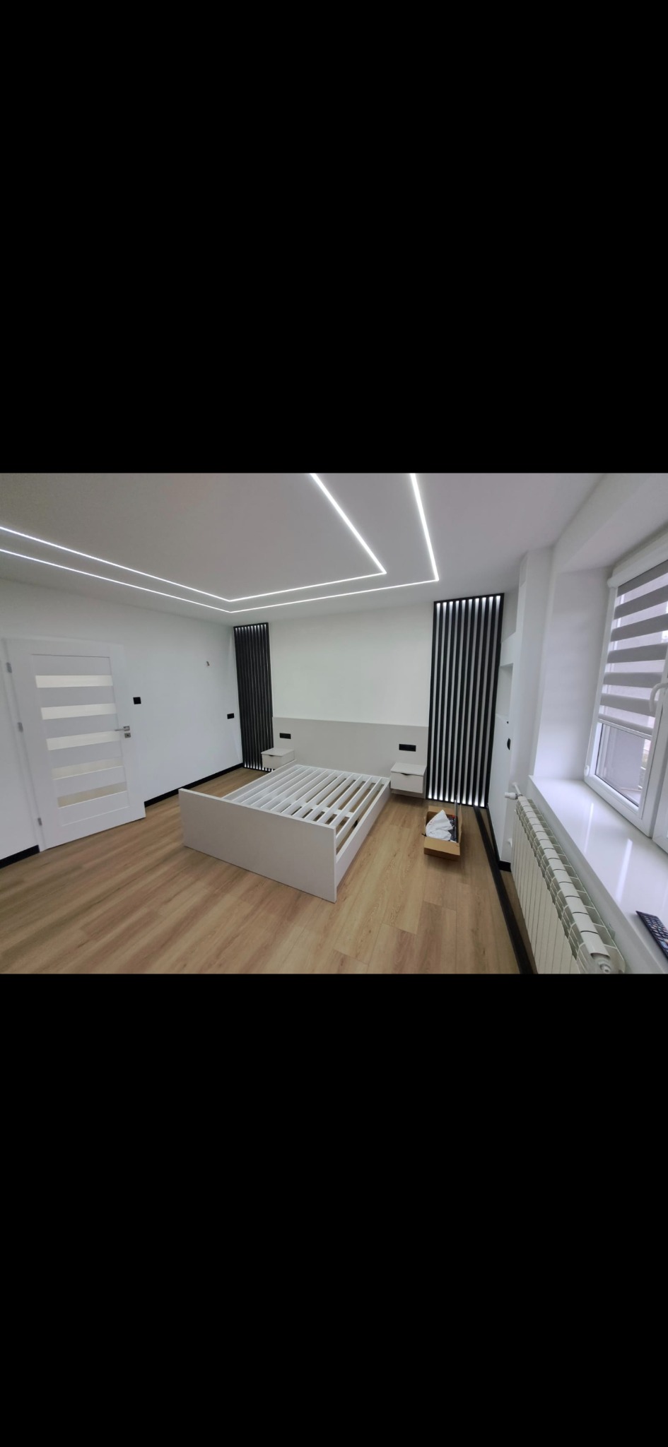 Biała sypialnia po remoncie: drewniana podłoga, rama łóżka bez materaca, grzejnik pod oknem, czarne panele dekoracyjne i nowoczesne oświetlenie sufitowe.