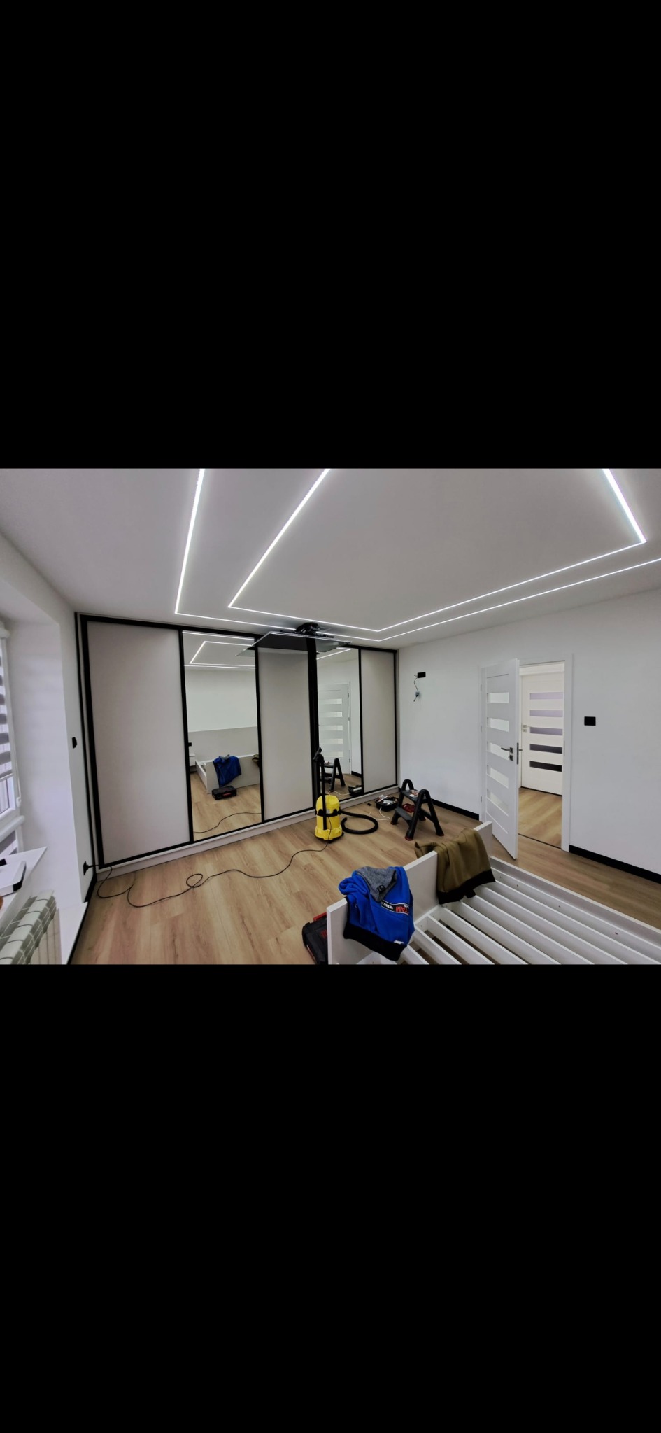 Nowoczesne wnętrze pokoju po remoncie: białe ściany, podłoga drewnopodobna, szafa z lustrzanymi drzwiami i geometryczne oświetlenie sufitowe LED.