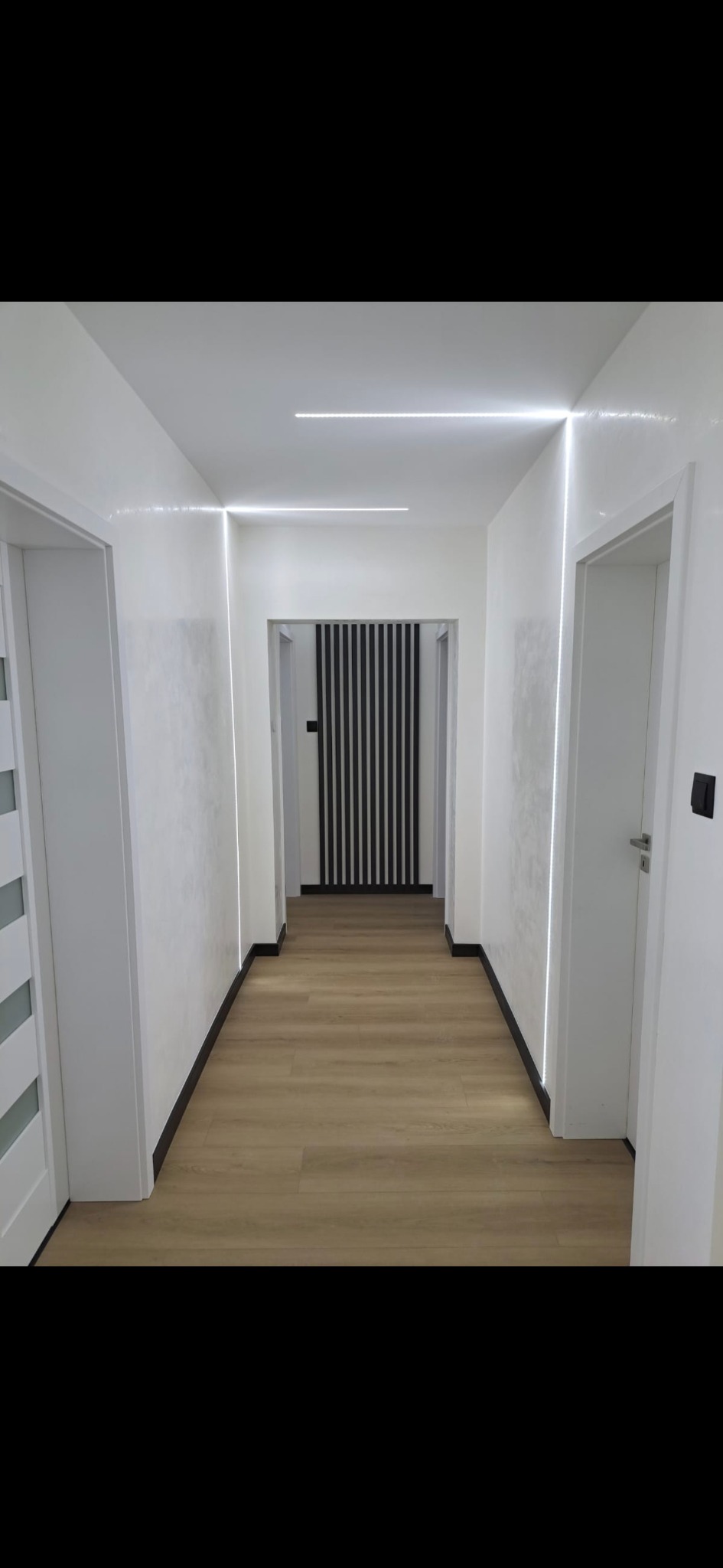 Nowoczesny korytarz z białymi ścianami, podświetleniem LED i panelami podłogowymi. Widoczne drzwi i dekoracyjna ścianka z czarnych listew w głębi.