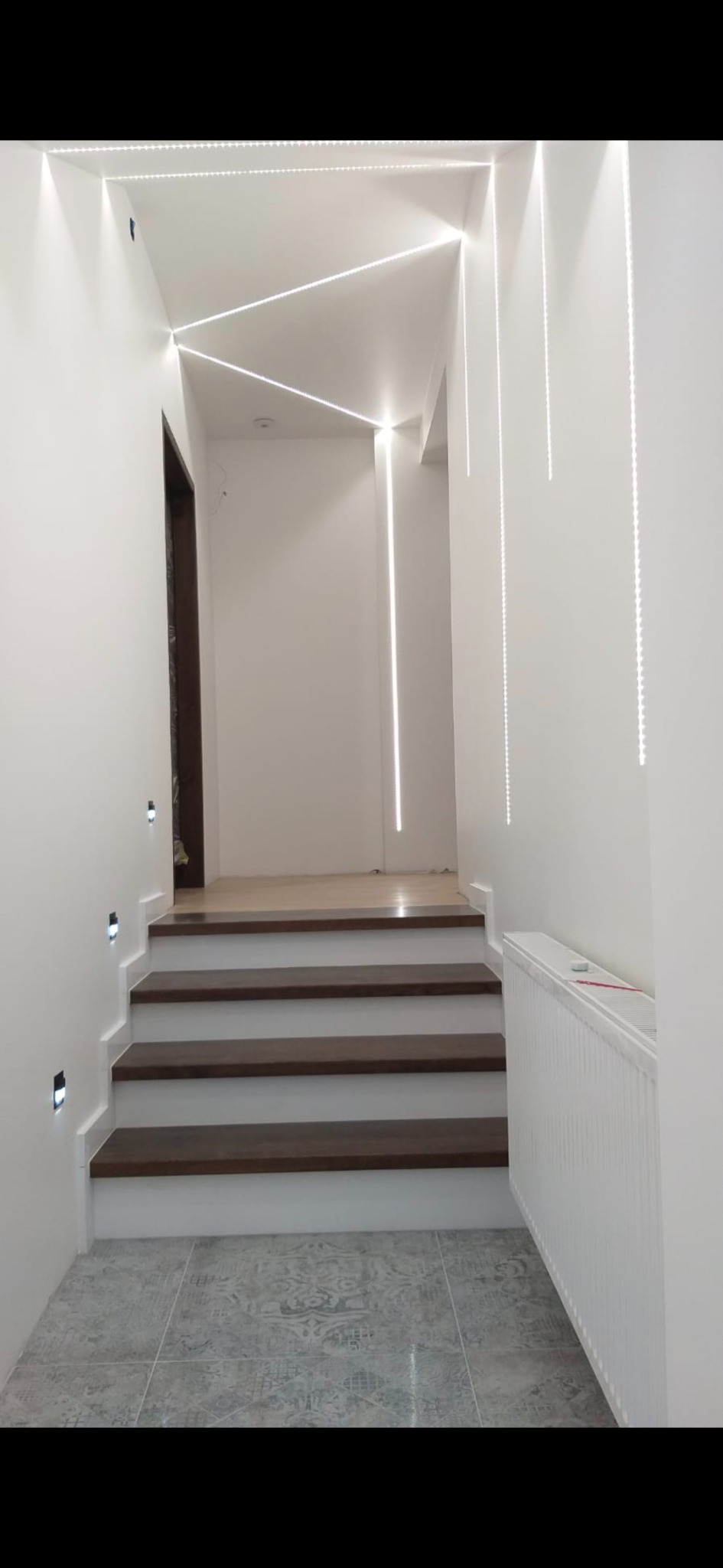 Nowoczesne oświetlenie LED podkreśla schody z ciemnego drewna i jasne ściany. Geometryczne wzory świetlne na suficie i pionowe linie na ścianach. Minimalistyczny design.