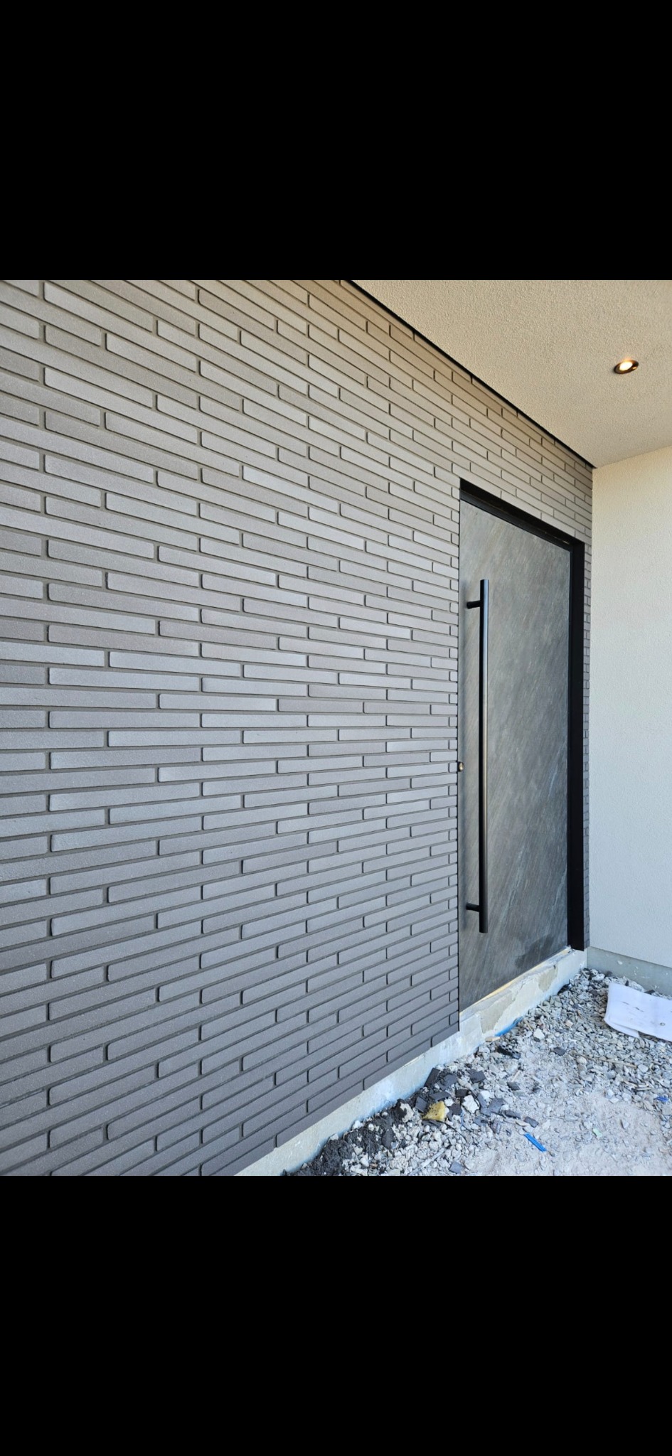 Elewacja budynku z szarej cegły klinkierowej i nowoczesnymi drzwiami wejściowymi z długim, czarnym uchwytem. Widoczne drobne prace wykończeniowe przy fundamencie.
