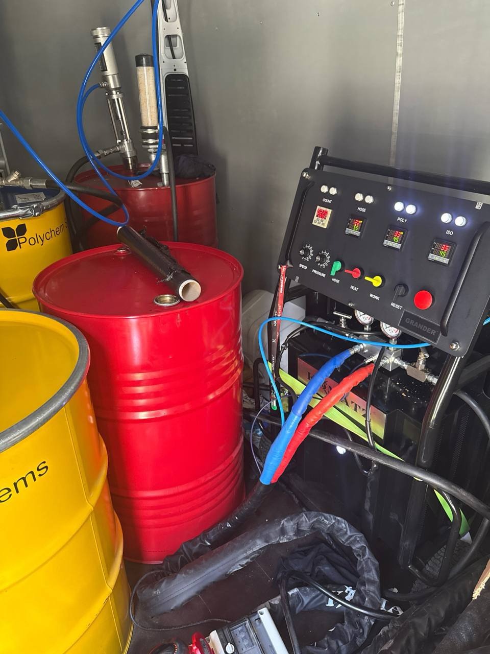 Mobilna jednostka natryskowa pianki PUR: beczki z komponentami, panel sterowania Grandera, węże i pistolet natryskowy w przestrzeni roboczej.