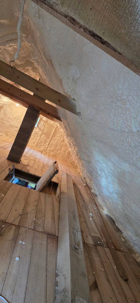 Poddasze z drewnianą podłogą i konstrukcją, pokryte jasną pianką izolacyjną, widoczne okno. Izolacja natryskowa w trakcie prac, surowe wykończenie.