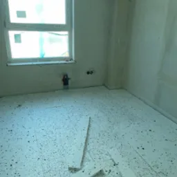 Przygotowanie podłoża: białe płyty izolacyjne z czarnym granulatem ułożone na podłodze w surowym wnętrzu z oknem. Widoczne instalacje i ślady prac na ścianach.