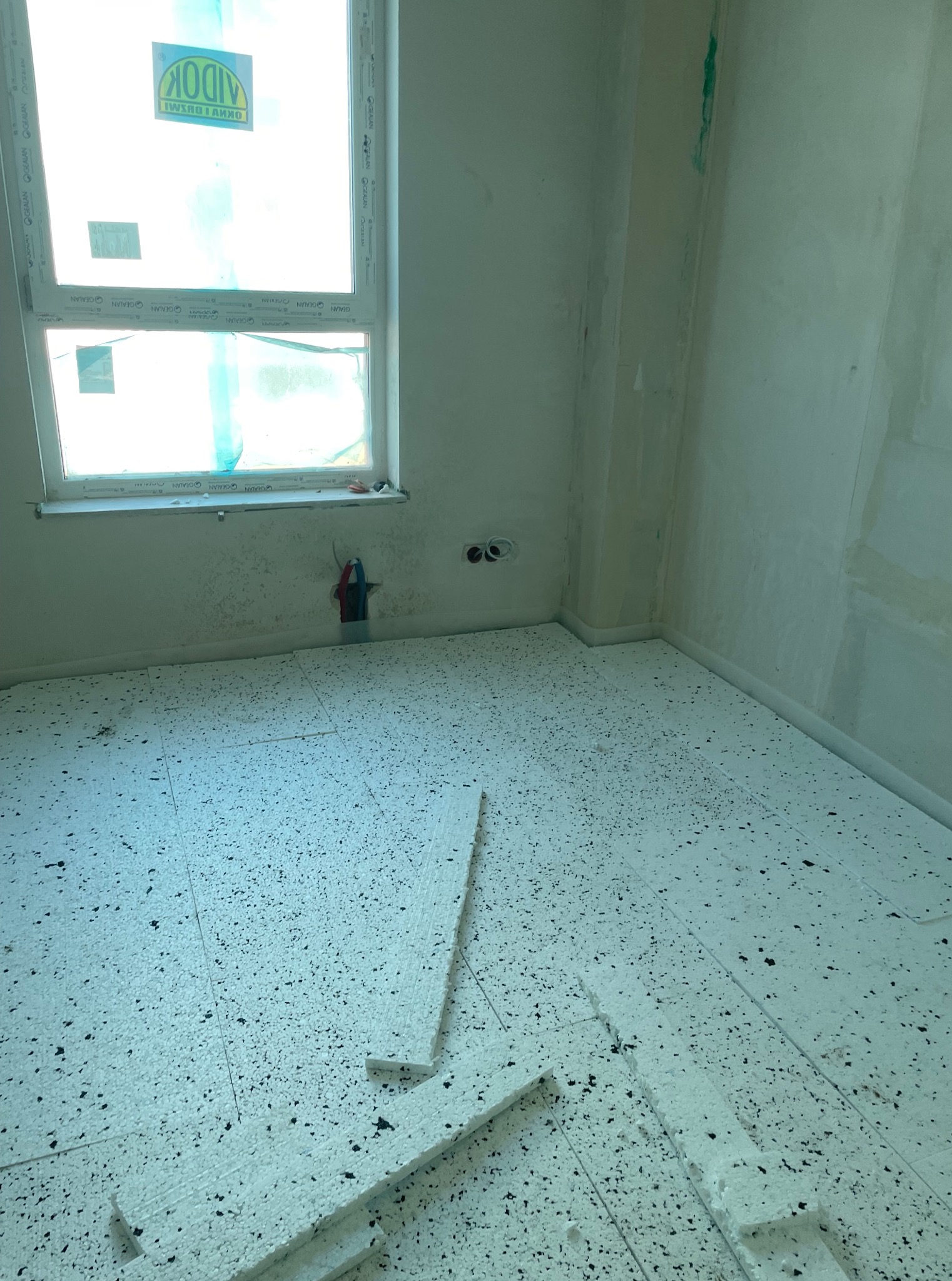 Przygotowanie podłoża: białe płyty izolacyjne z czarnym granulatem ułożone na podłodze w surowym wnętrzu z oknem. Widoczne instalacje i ślady prac na ścianach.