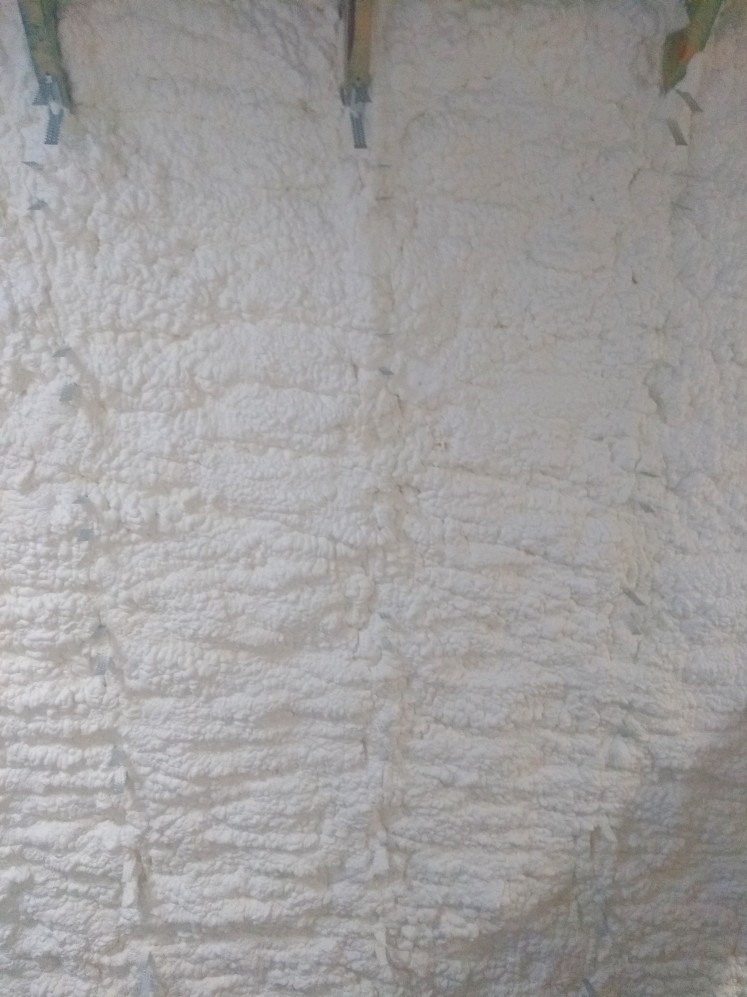 Poddasze pokryte białą, gęstą pianką izolacyjną, widoczne elementy konstrukcyjne. Tekstura pianki nierówna, przypominająca strukturę chmur. Dobre ocieplenie.