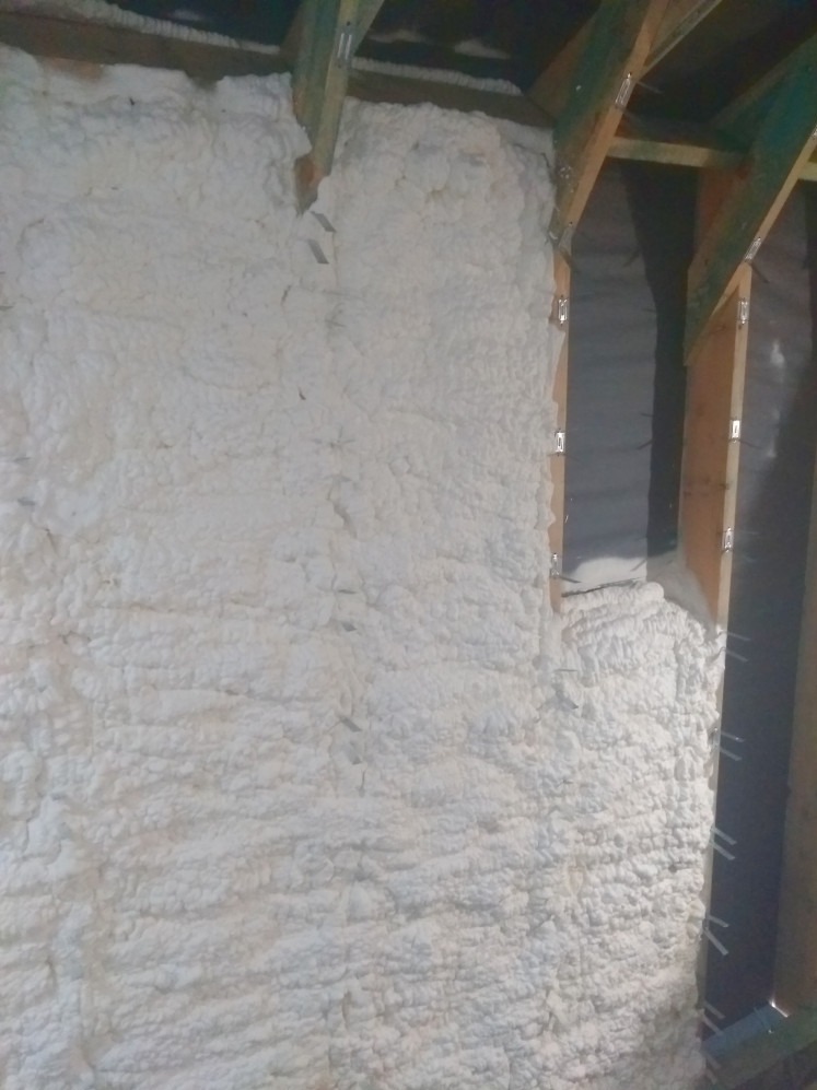 Ściana poddasza z drewnianą konstrukcją pokryta białą, nierównomierną warstwą pianki izolacyjnej, widoczne elementy mocujące. Izolacja natryskowa.