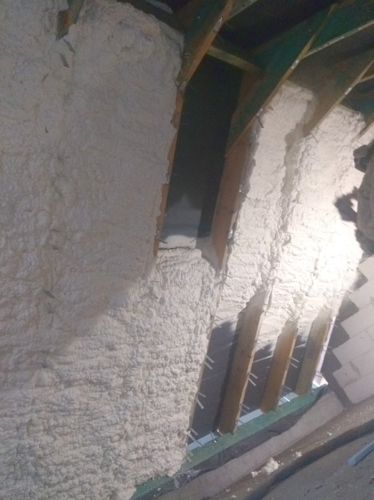 Poddasze z drewnianą konstrukcją ocieplone białą pianką PUR, widoczne szpilki mocujące. Oświetlenie boczne podkreśla nierównomierną strukturę piany.