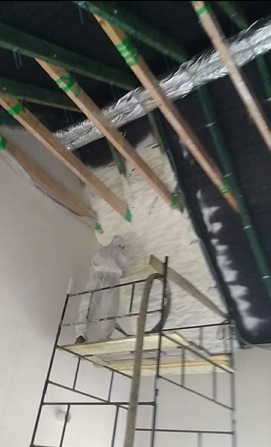 Ocieplanie poddasza pianką: pracownik w kombinezonie na rusztowaniu aplikuje izolację między krokwiami. Widoczna folia paroizolacyjna i elementy konstrukcji dachu.