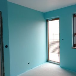 Bartłomiej Skonieczny - Pokój po malowaniu na odcień błękitu, z widocznymi oknami i fragmentem futryny drzwiowej w trakcie wykończenia. Podłoga zabezpieczona folią.
