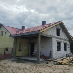 Bartłomiej Skonieczny - Dom w trakcie rozbudowy: część pomalowana na żółto, druga z białego bloczka, czerwona dachówka, widoczne rusztowania i materiały budowlane. Pochmurne niebo w tle.