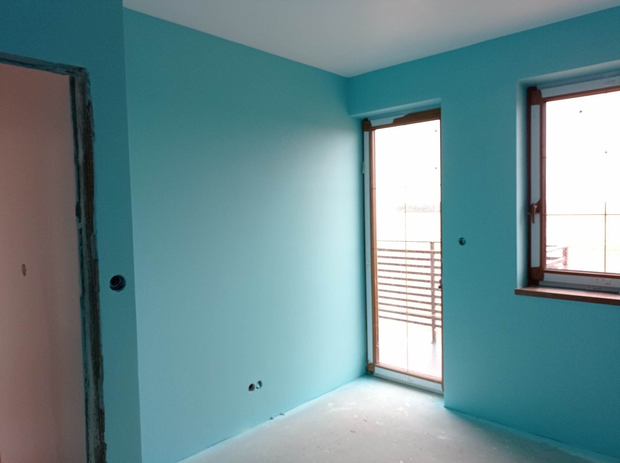 Pokój po malowaniu na odcień błękitu, z widocznymi oknami i fragmentem futryny drzwiowej w trakcie wykończenia. Podłoga zabezpieczona folią.