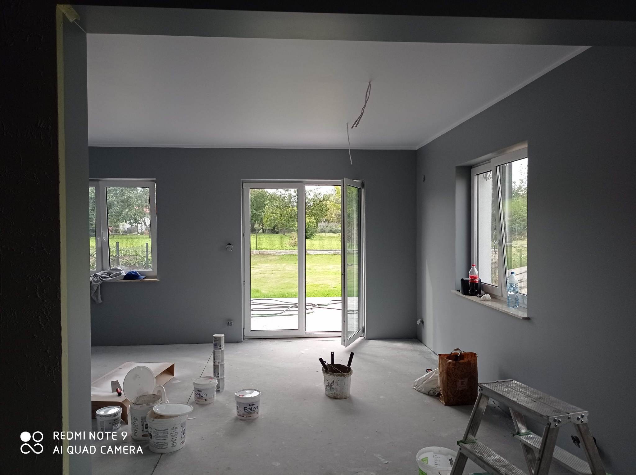 Pomieszczenie po malowaniu na szaro z białym sufitem, widoczne okna, drzwi balkonowe, drabina i puszki z farbą. Kabel zwisa z sufitu.