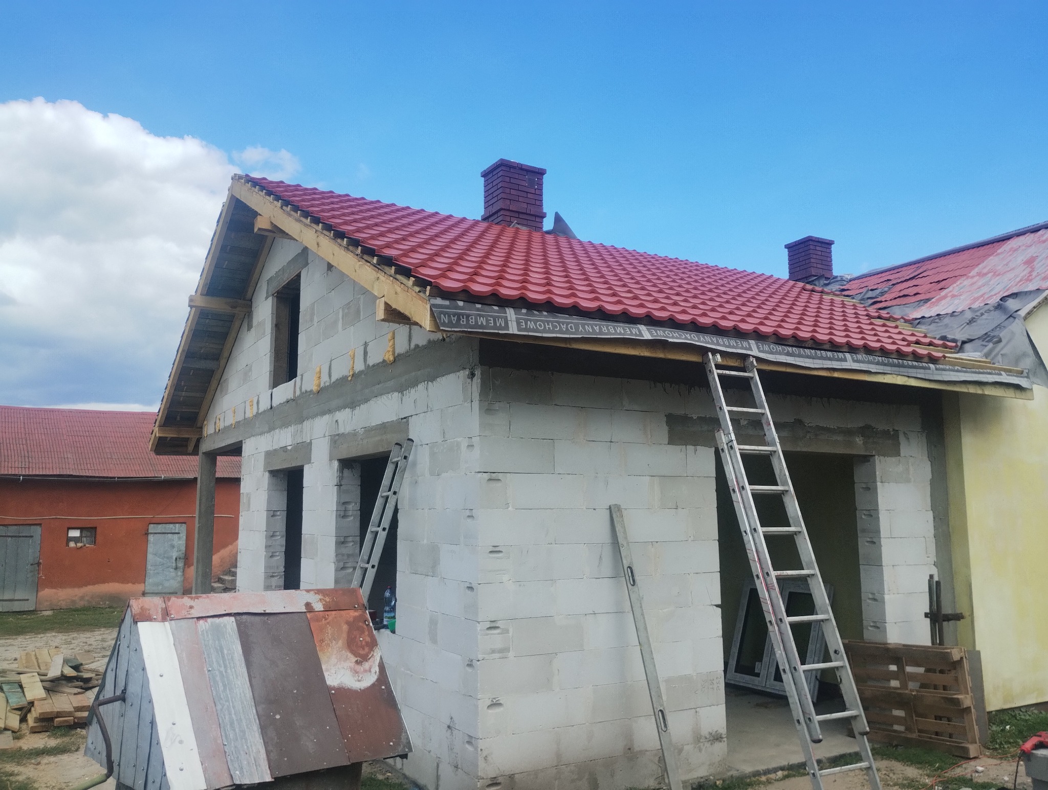 Budowa domu z białych bloczków: czerwony dach, okna bez szyb, drabina oparta o ścianę. Widoczne materiały budowlane i fragment sąsiedniego budynku.