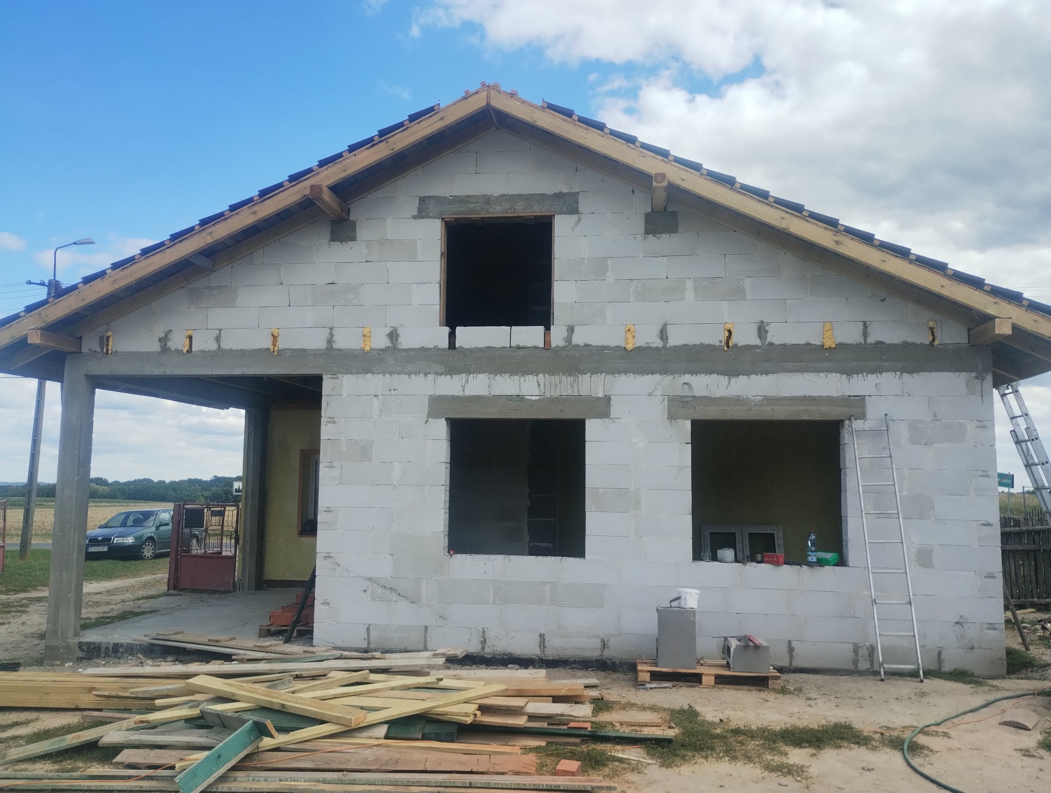 Budowa domu z betonu komórkowego w trakcie realizacji. Widoczne niezadaszone okna, drewniane elementy konstrukcji dachu i porozrzucane deski.