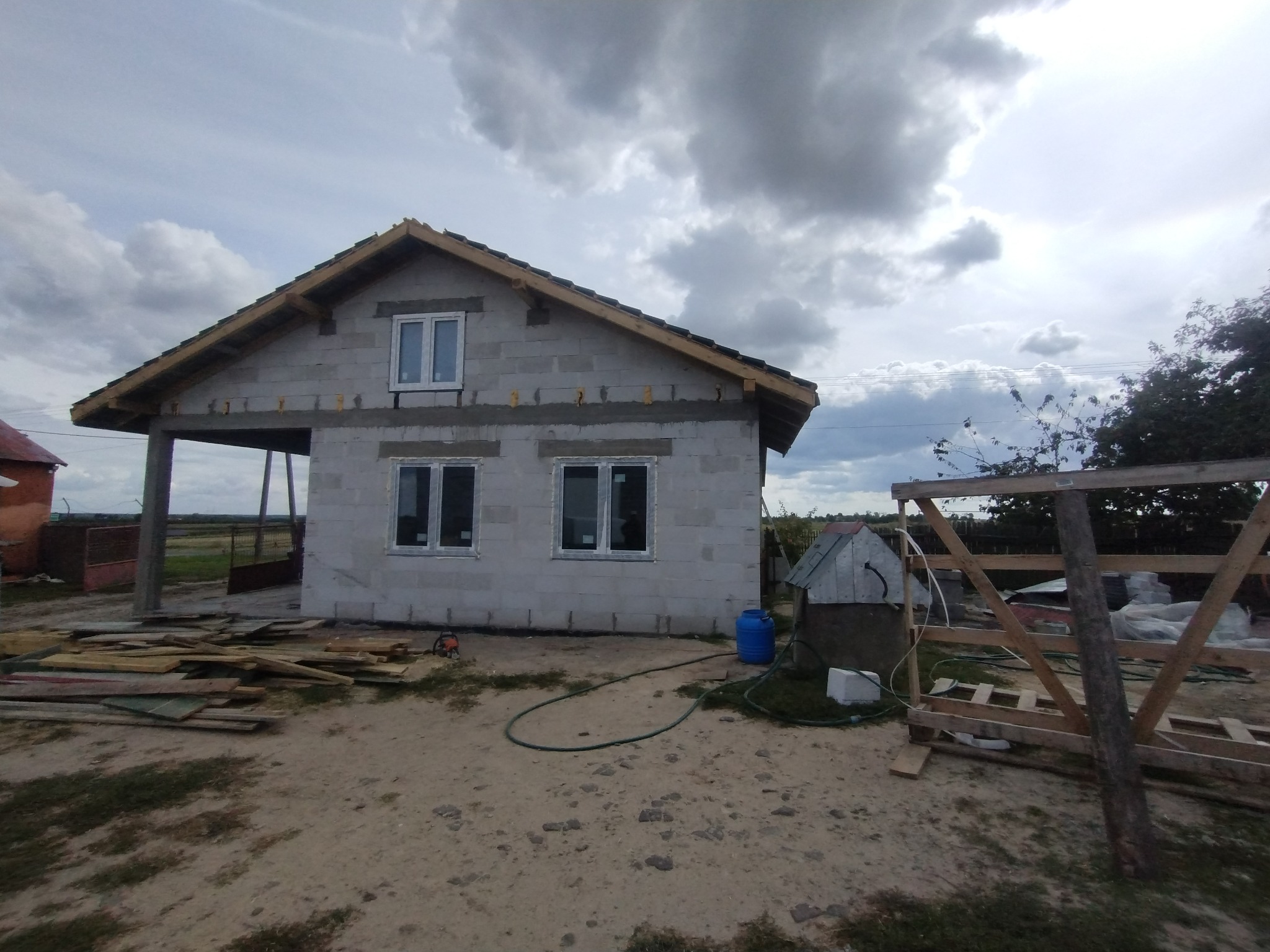 Surowy budynek jednorodzinny w trakcie budowy, z oknami i drewnianą konstrukcją dachu, otoczony placem budowy z materiałami i narzędziami.