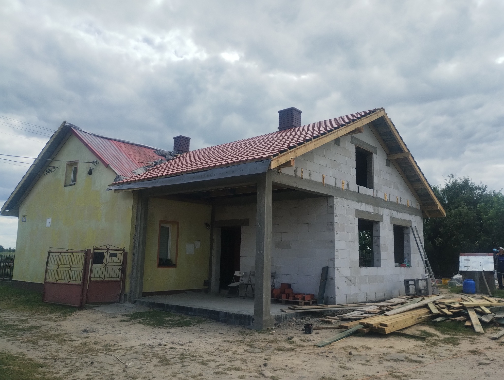 Dom w trakcie rozbudowy: część pomalowana na żółto, druga z białego bloczka, czerwona dachówka, widoczne rusztowania i materiały budowlane. Pochmurne niebo w tle.