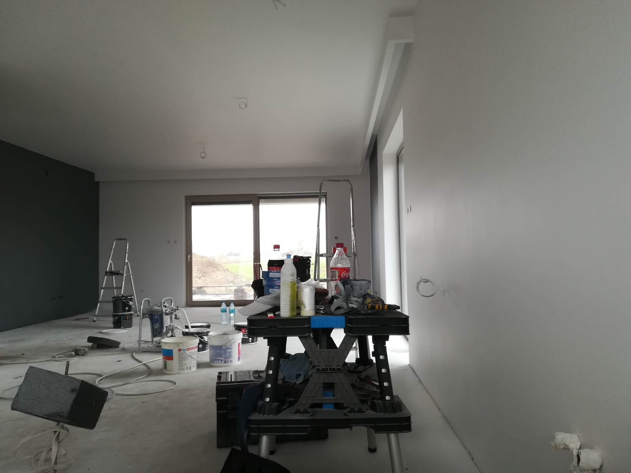 Wnętrze w trakcie malowania: ściany w szarym odcieniu, narzędzia malarskie na stole warsztatowym, drabina w tle. Widoczne niedoskonałości na ścianach przed ostatecznym wykończeniem.