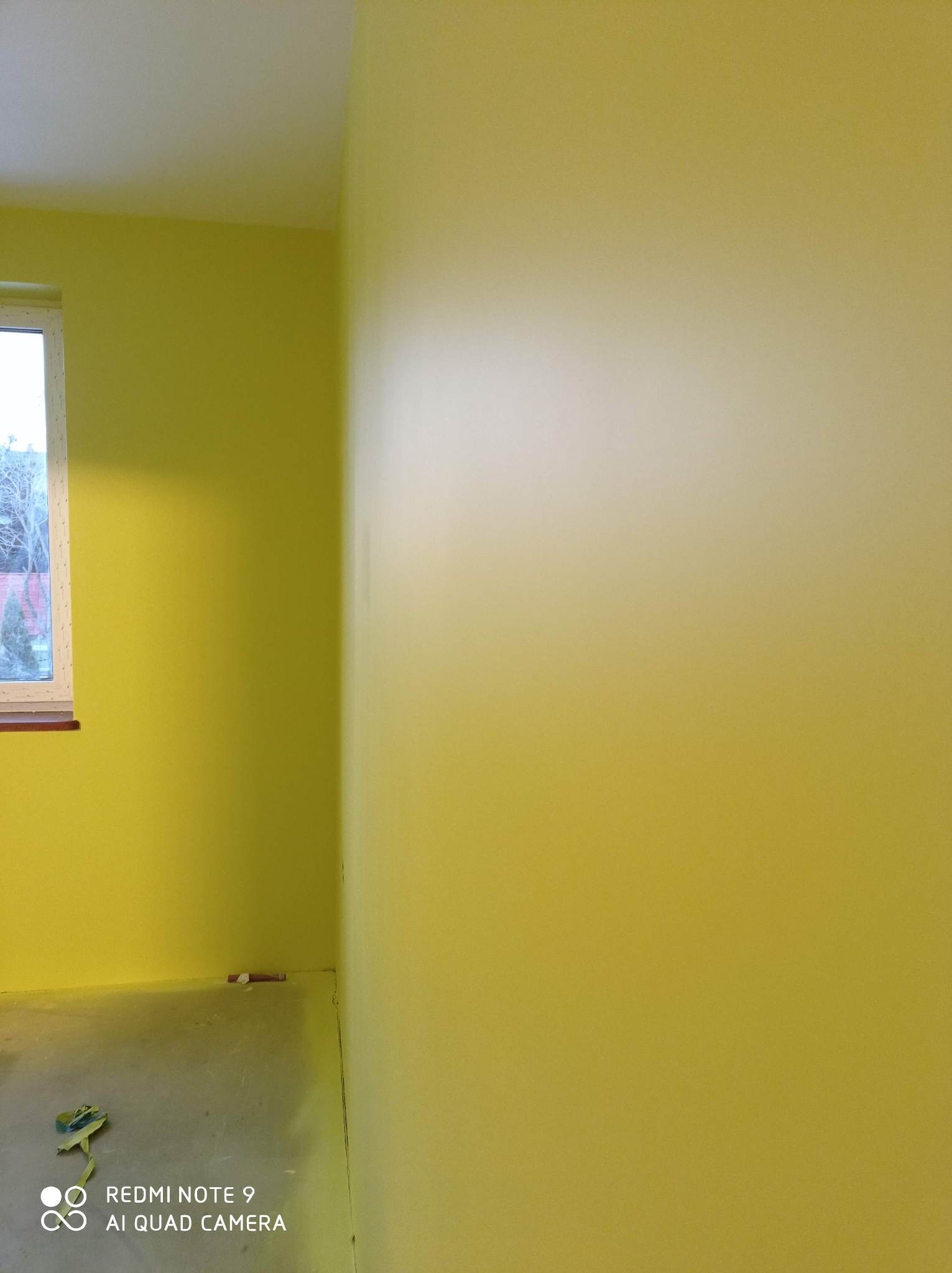 Ściana pomalowana na żółto w narożniku pokoju z oknem. Widoczne ślady farby na podłodze i taśma malarska. Świeżo pomalowane ściany w jasnym odcieniu żółci.