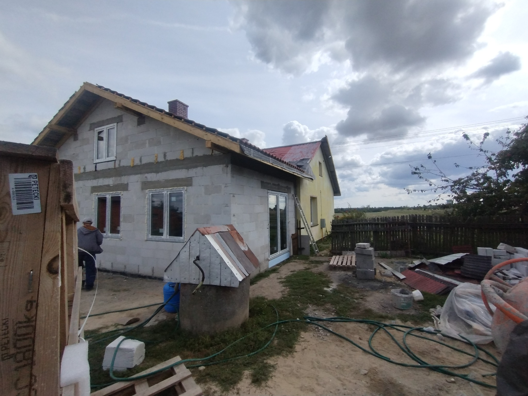 Budowa domu jednorodzinnego w trakcie realizacji: surowe ściany z pustaków, zamontowane okna, widoczna studnia i materiały budowlane na posesji.