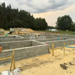 Fundamenty domu z bloczków betonowych na piaszczystym terenie. Widoczne zbrojenia, niebieska rura. W tle las i sprzęt budowlany.