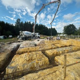 Tecton sp. z o. o. - Wylewanie betonu z pompy do wykopów zbrojeniowych pod fundamenty domu na działce z lasem w tle, widoczna betoniarka i pracownicy w białych kombinezonach.