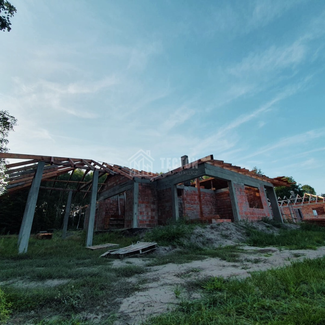 Wznoszenie domu z czerwonej cegły: surowe ściany, betonowe słupy i drewniana konstrukcja dachu na tle błękitnego nieba. Widok z poziomu gruntu, wczesny etap budowy.