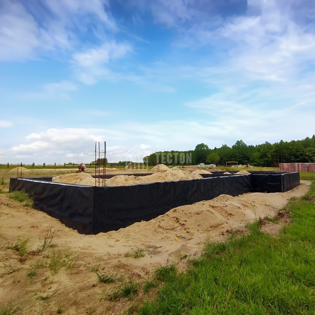 Fundament domu w trakcie budowy, zabezpieczony czarną folią, z widocznym zbrojeniem i nasypanym piaskiem. W tle zielone drzewa i błękitne niebo.