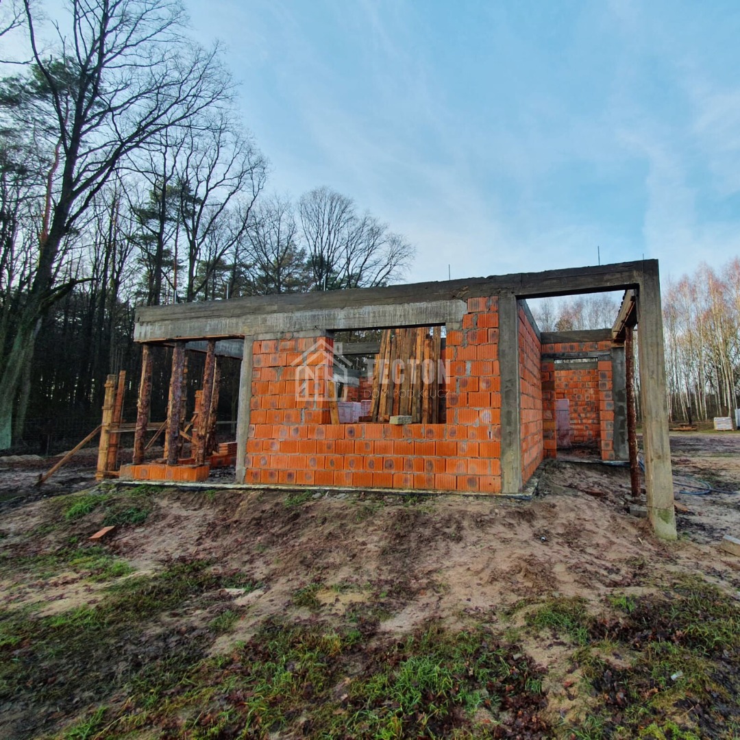 Surowy stan domu z czerwonej cegły z betonowymi nadprożami, na tle lasu i błękitnego nieba. Widoczne drewniane podpory i elementy konstrukcyjne.