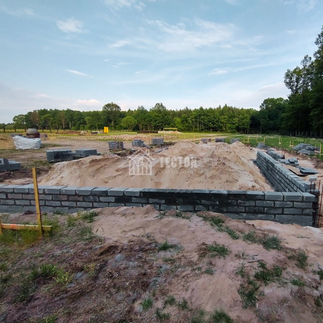 Fundamenty domu z szarych bloczków betonowych na piaszczystym terenie, w tle zieleń drzew i błękitne niebo. Widoczne zbrojenia i przygotowany teren pod budowę.