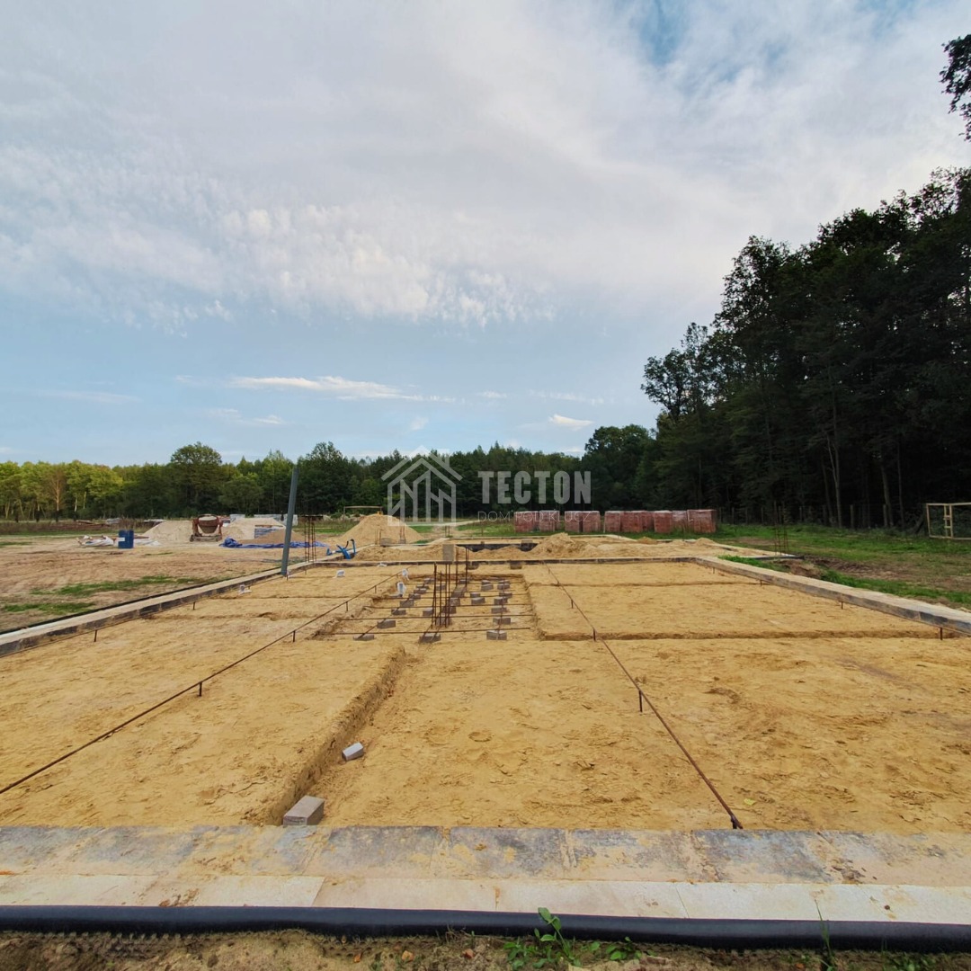 Fundamenty domu zbrojone stalą, przygotowane do zalania betonem. Widoczne szalunki, teren budowy i las w tle. Ujęcie z szerokim kątem.