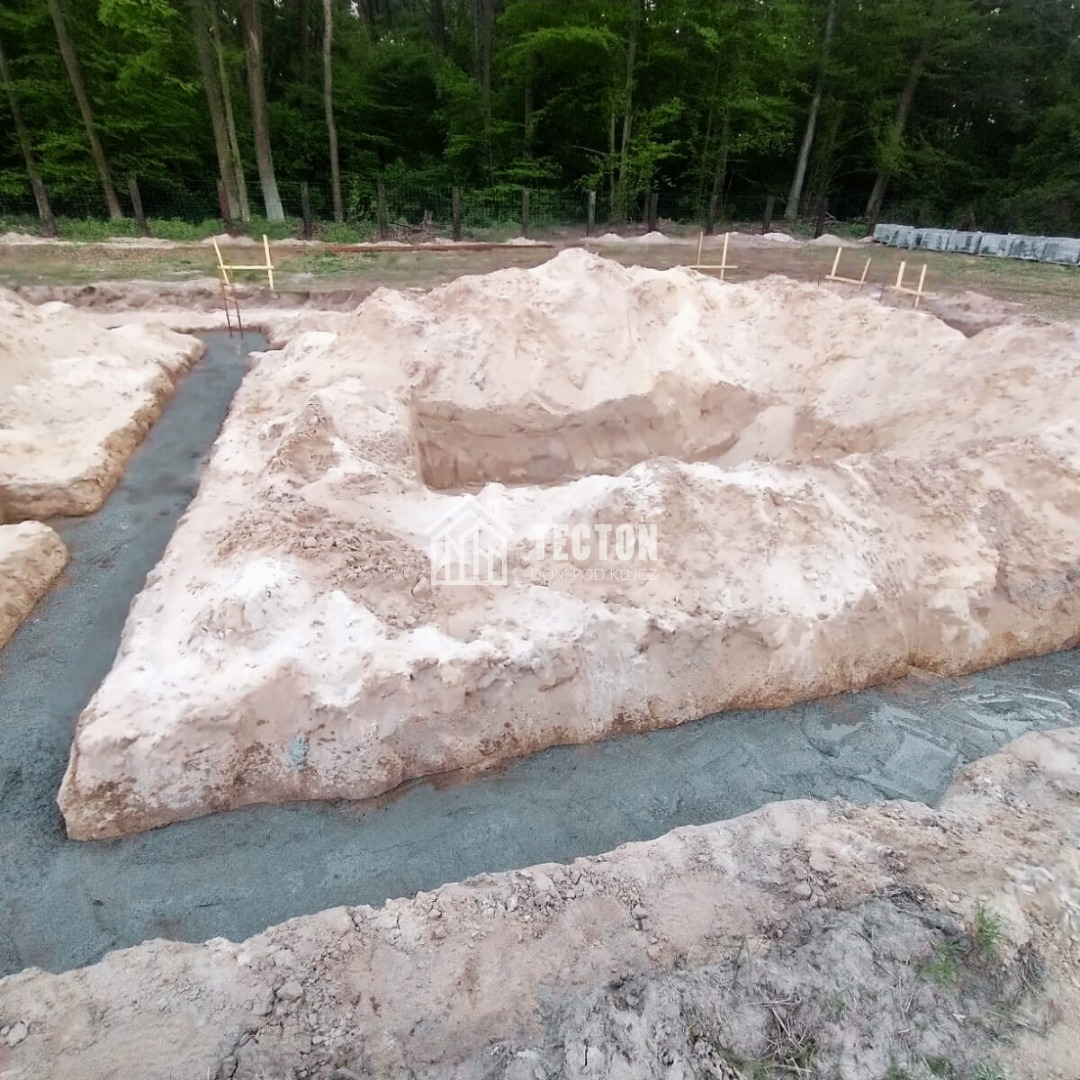 Wykop pod fundamenty domu z częściowo wylaną ławą fundamentową, widoczne drewniane znaczniki, tło z drzew. Ujęcie z góry.