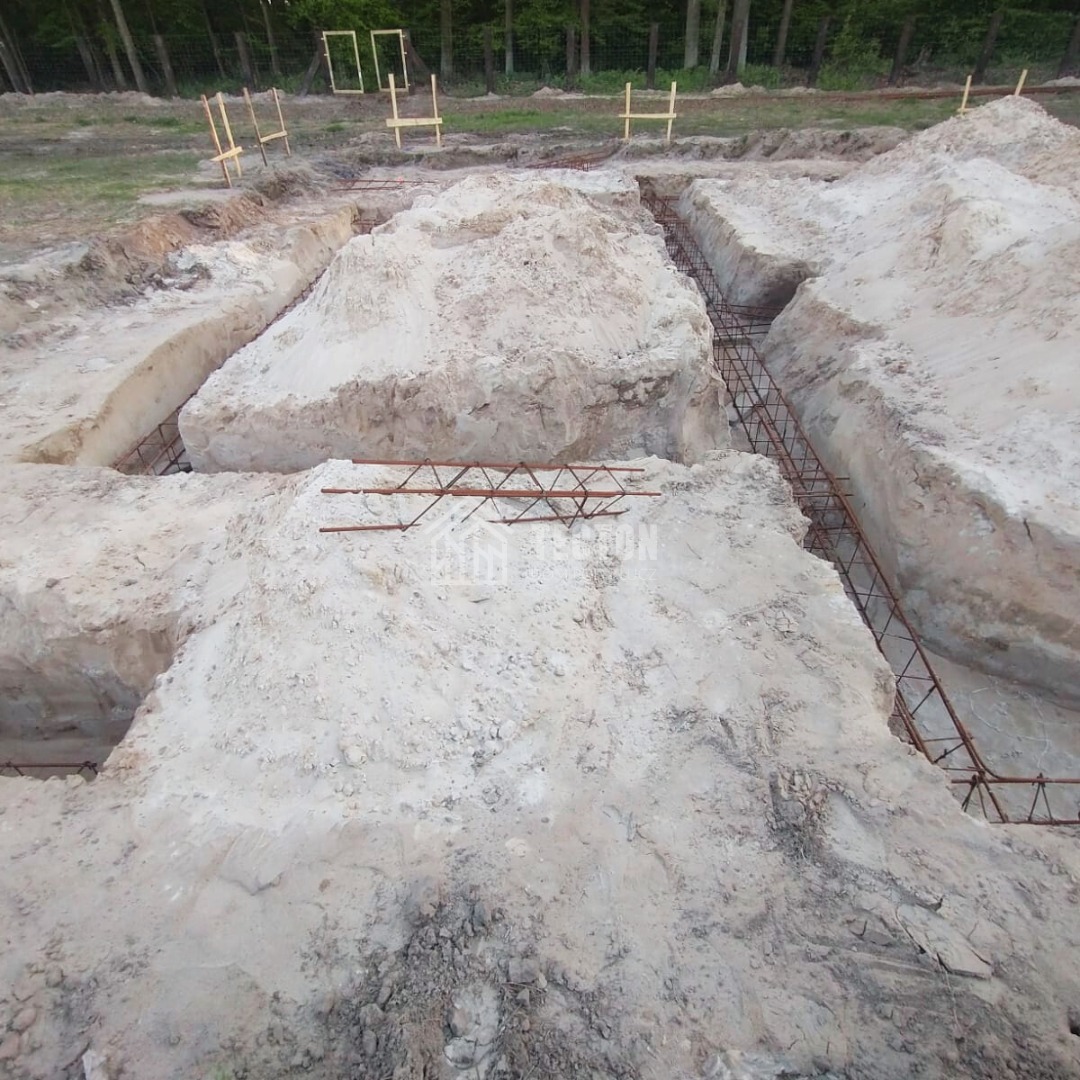Zbrojenie fundamentów w wykopach, przygotowane pod wylanie betonu. Widoczne drewniane szalunki i ukształtowany teren pod budowę domu.