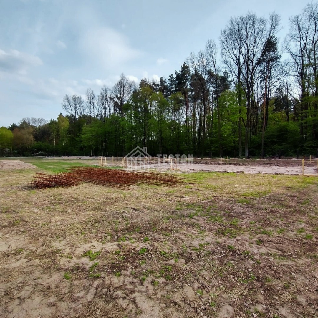 Przygotowanie terenu pod budowę domu: rozłożone zbrojenie na gruncie, wytyczone fundamenty, las w tle. Widok z poziomu gruntu.