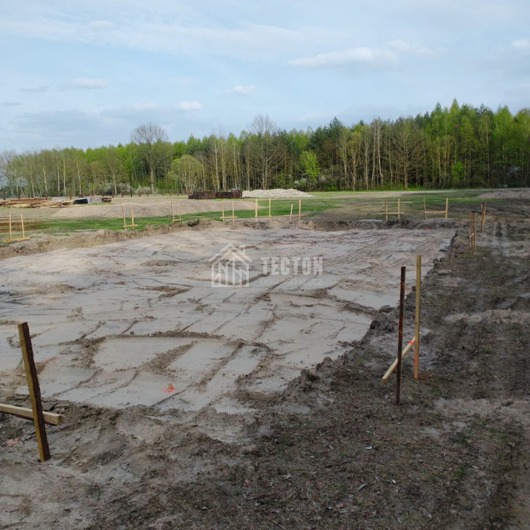 Przygotowany teren pod budowę domu parterowego z wytyczonym obrysem fundamentów, widoczne drewniane paliki i logo firmy Tecton w tle las.