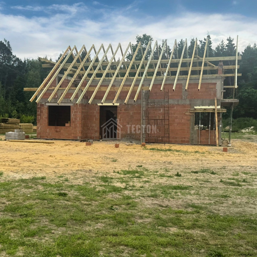 Murowany dom w trakcie budowy: ściany z czerwonej cegły, drewniana konstrukcja dachu, rusztowanie, stos desek w tle, zielona trawa i las w tle.