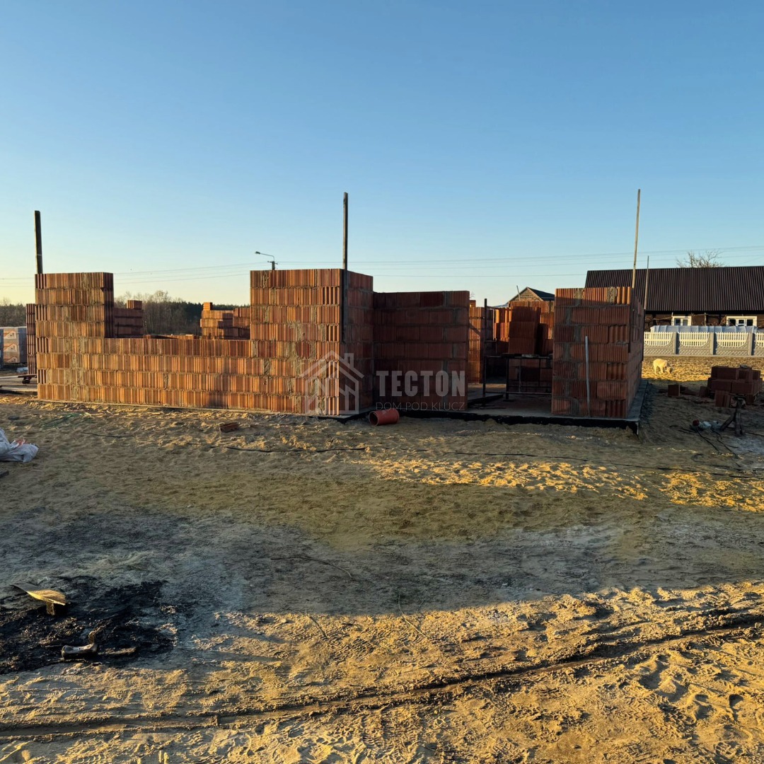 Wznoszenie ścian z czerwonej cegły na fundamencie, widoczne zbrojenie i logo firmy Tecton Dom pod klucz. W tle zabudowa jednorodzinna i błękitne niebo.