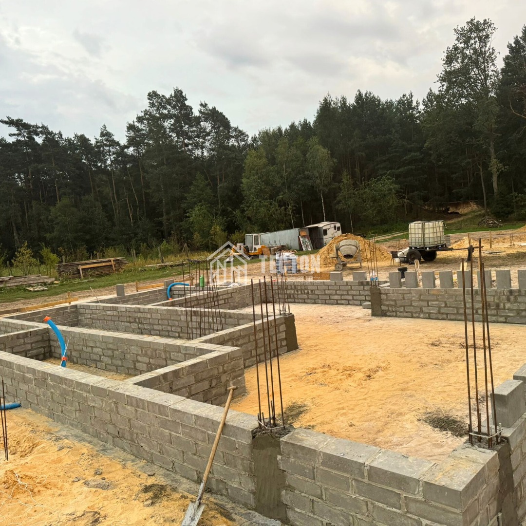 Wznoszenie fundamentów z bloczków betonowych na piaszczystym podłożu. Widoczne wystające pręty zbrojeniowe, narzędzia i otoczenie budowy. Etap początkowy budowy domu.