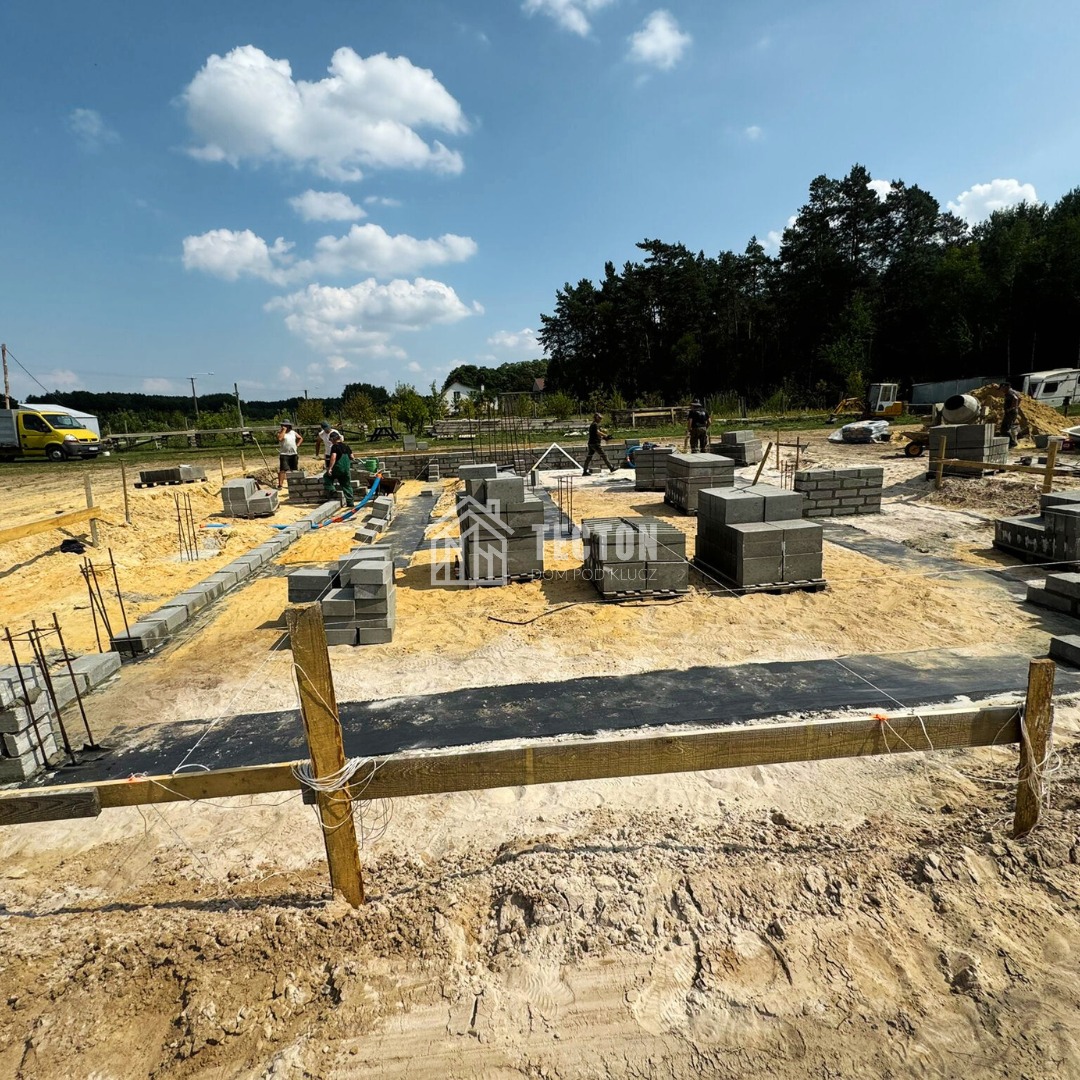 Budowa fundamentów domu z bloczków betonowych na piaszczystym terenie. Widoczni pracownicy i sprzęt budowlany w słoneczny dzień.