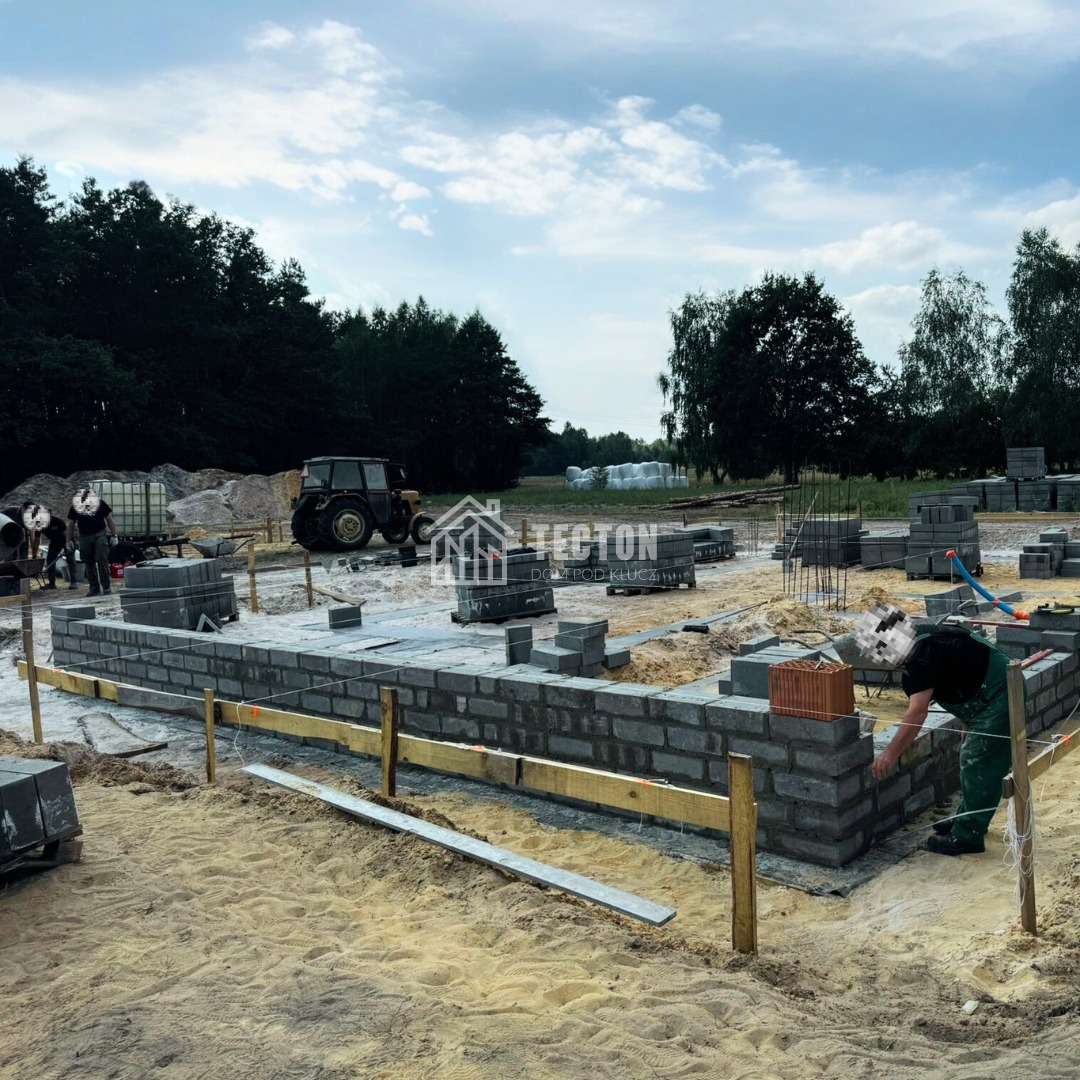 Budowa fundamentów domu z bloczków betonowych, widoczny pracownik w kombinezonie, teren budowy z traktorem i zapleczem, las w tle, pochmurne niebo.
