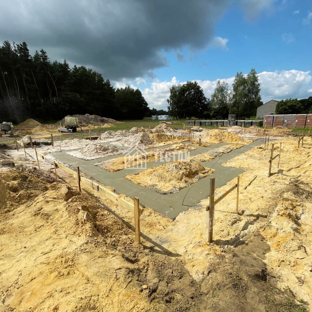 Fundamenty wylane betonem, drewniane szalunki, widok na plac budowy domu w słoneczny dzień. W tle drzewa i niebo z chmurami.