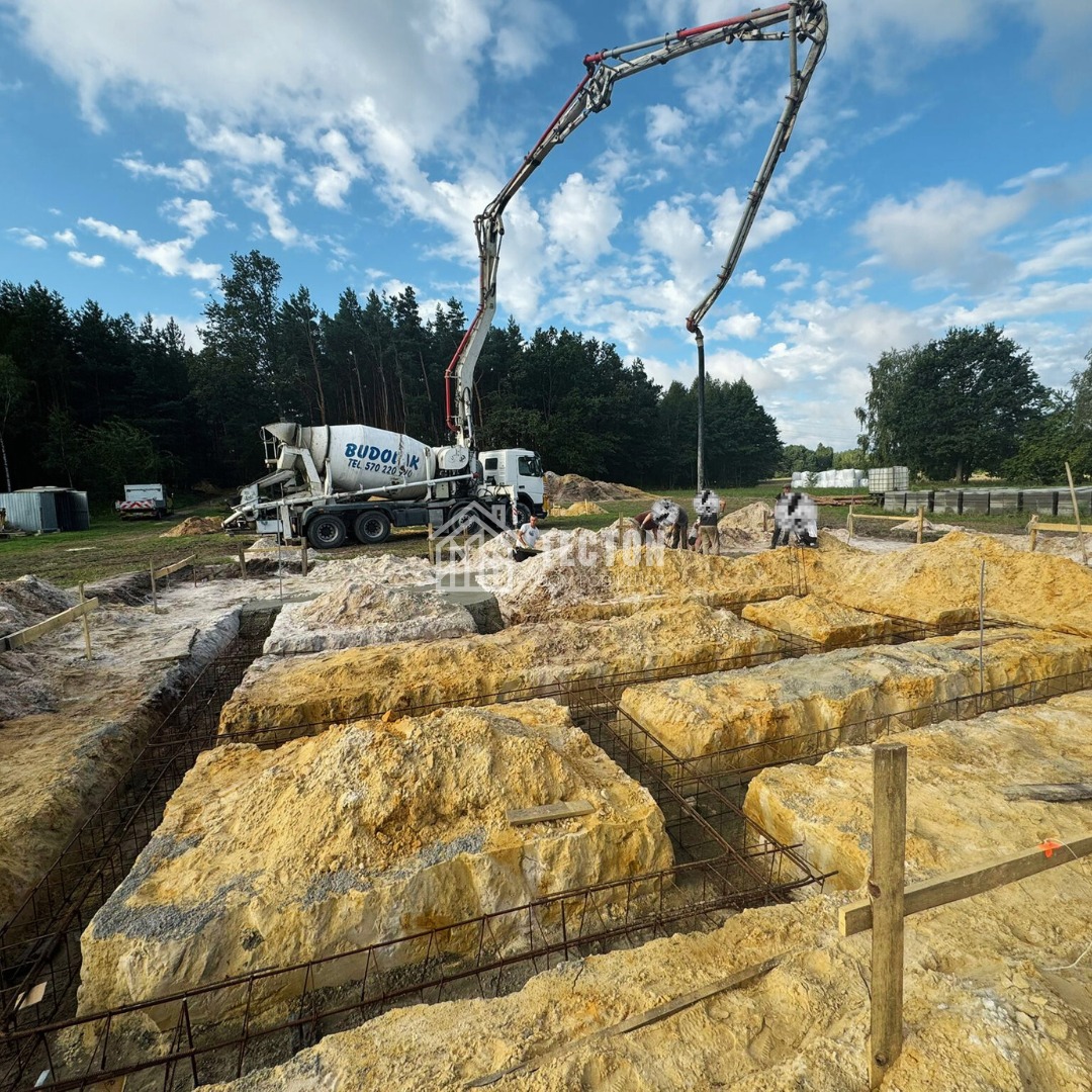 Wylewanie betonu z pompy do wykopów zbrojeniowych pod fundamenty domu na działce z lasem w tle, widoczna betoniarka i pracownicy w białych kombinezonach.