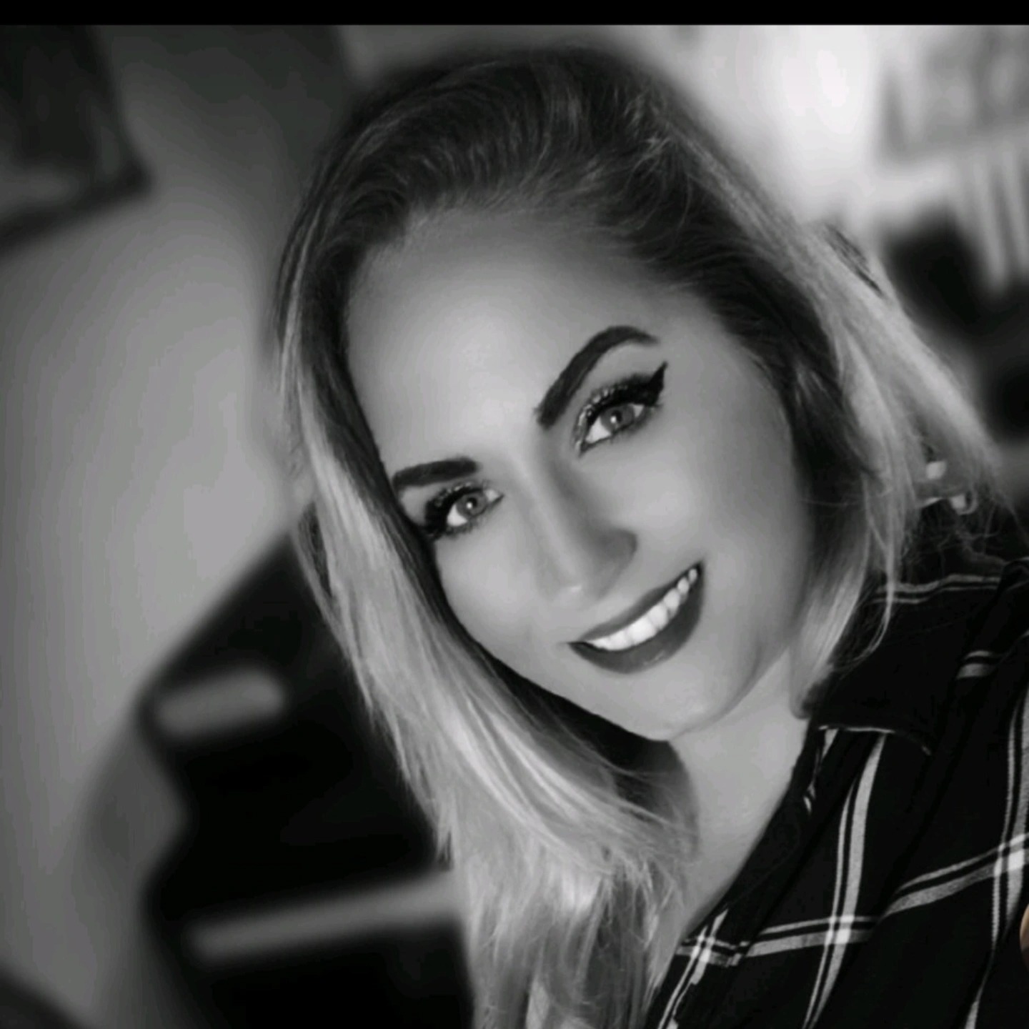 Portret uśmiechniętej kobiety w czarno-białej tonacji, z wyrazistym makijażem oczu i ciemną szminką, ubrana w koszulę w kratę.