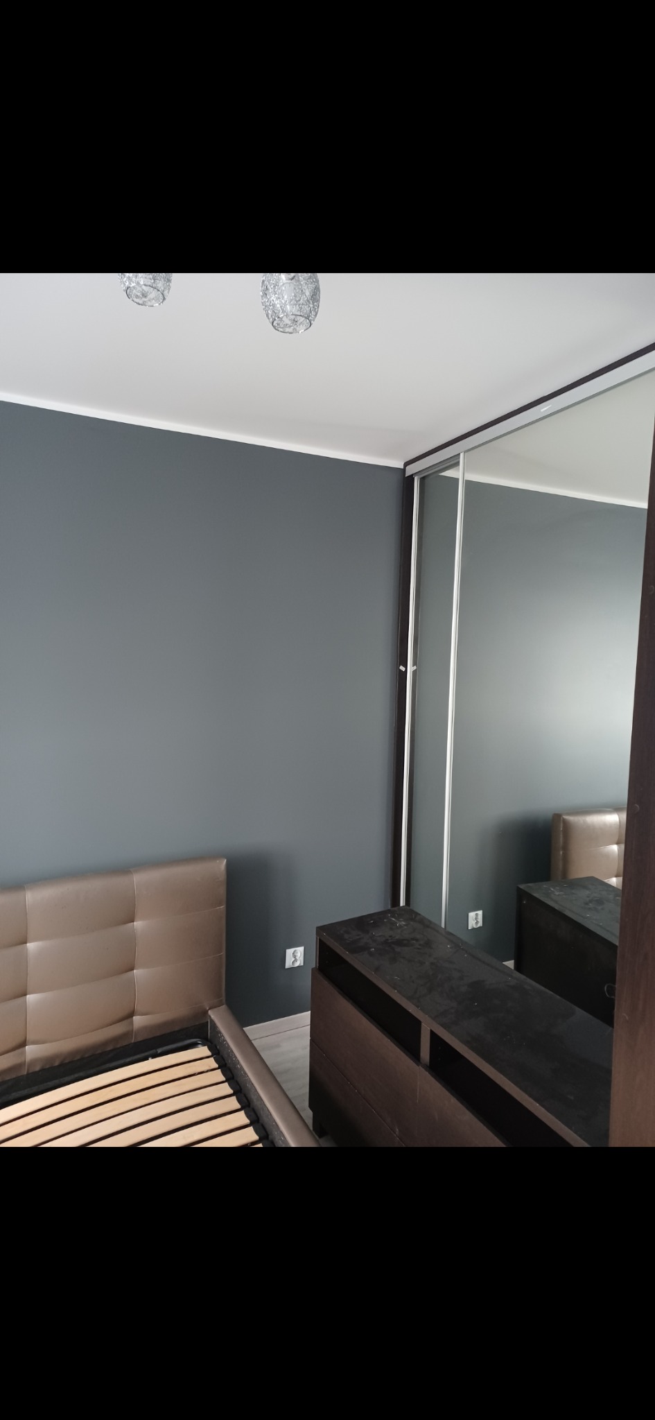 Ściana w odcieniach szarości po malowaniu w sypialni, widoczny fragment łóżka i szafa przesuwna z lustrami. Minimalistyczny design, eleganckie wykończenie.