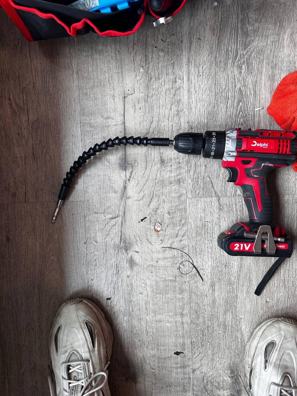 Wiertarko-wkrętarka Delphi 21V z elastycznym przedłużeniem bitu na podłodze imitującej drewno, obok widoczne buty i torba narzędziowa.