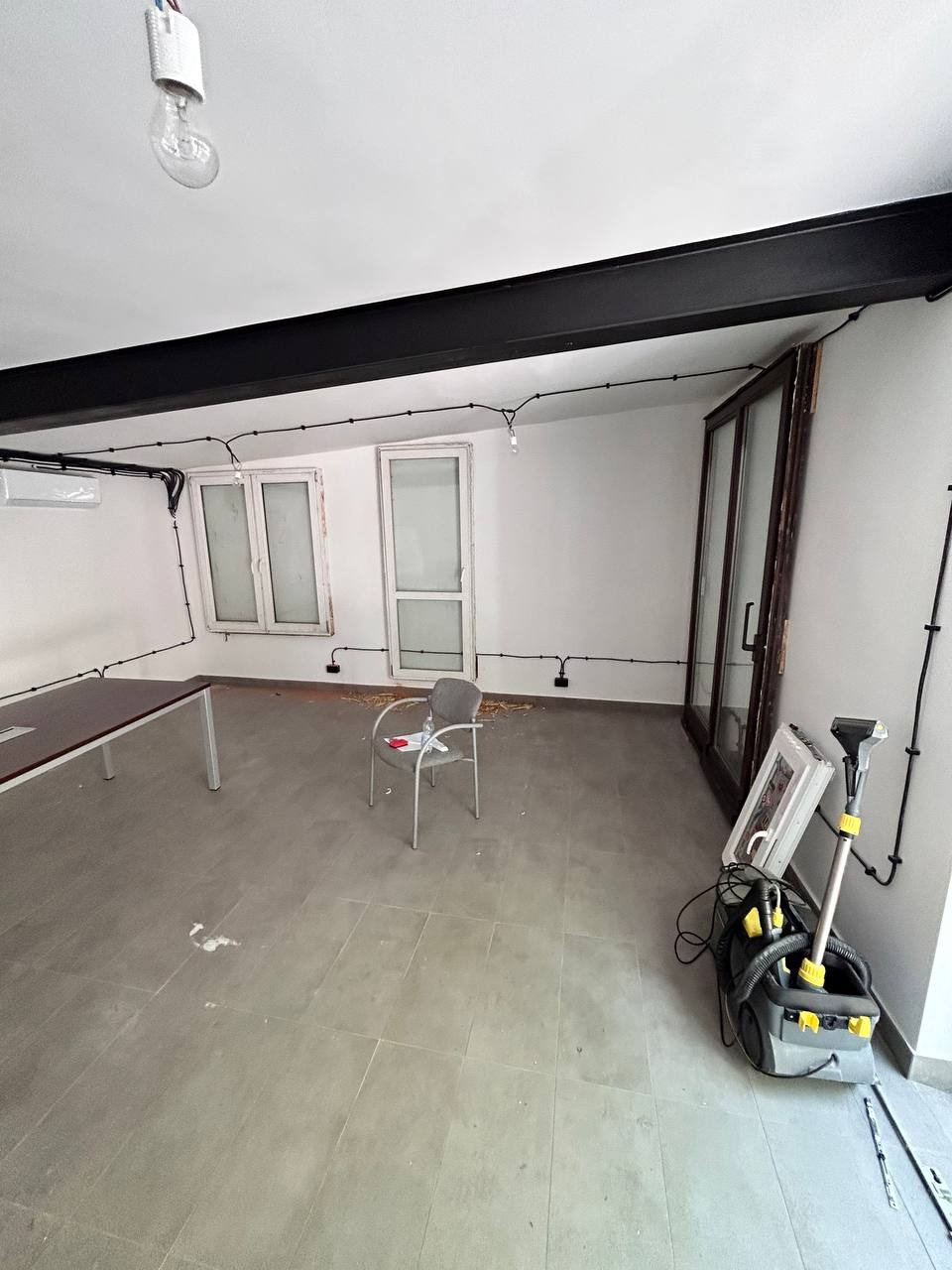 Wnętrze po remoncie: widoczne okna z białymi ramami, przesuwne drzwi balkonowe, szara podłoga z płytkami i odkurzacz do czyszczenia dywanów. Minimalistyczny wystrój, surowy beton.